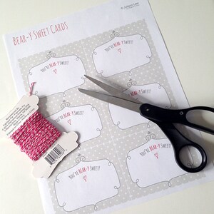 May include: A printable sheet of paper with five bear-themed gift tags. The tags are decorated with a polka dot pattern and the text "You're Bear-y Sweet!" with a small heart. A pair of scissors and a spool of pink and white twine are also in the image.