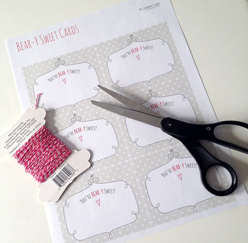 May include: A printable sheet of paper with five bear-themed gift tags. The tags are decorated with a polka dot pattern and the text "You're Bear-y Sweet!" with a small heart. A pair of scissors and a spool of pink and white twine are also in the image.