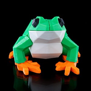 May include: A green and white geometric frog figurine with orange feet. The frog is made of plastic and has a stylized design.