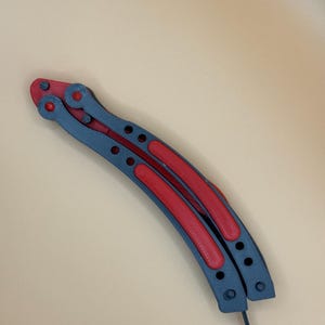 May include: A gray and red metal butterfly knife with a curved blade and a handle with holes.