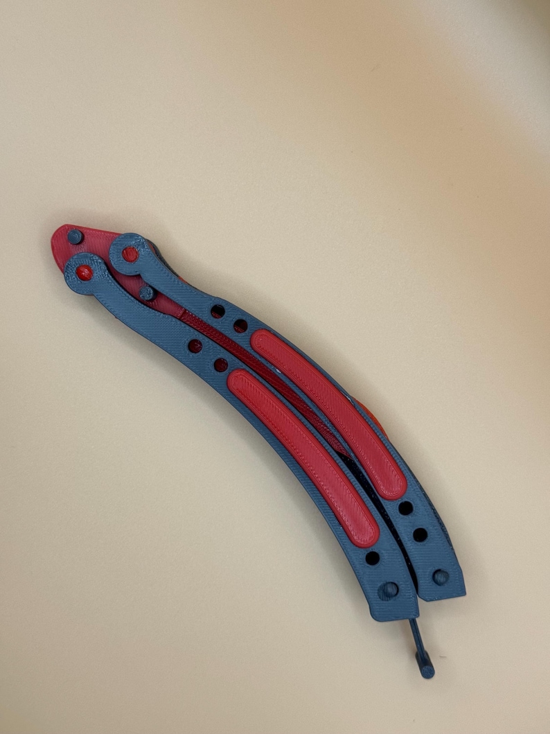 May include: A gray and red metal butterfly knife with a curved blade and a handle with holes.