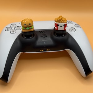 以下が含まれることがあります： 白いPlayStation 5 DualSenseコントローラーに、カスタムのサムスティックキャップが2つ取り付けられています。左側のキャップはハンバーガー、右側のキャップはフライドチキンが入った箱です。