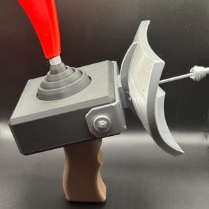 Puede incluir: Un mando de juego personalizado con un joystick rojo, una base gris y un mango marrón. El mando presenta un componente plateado en forma de escudo fijado en el lateral. El diseño es moderno y parece impreso en 3D.