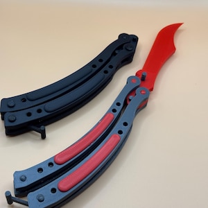 May include: Two butterfly knives, one black and one gray with red accents. The knives are folded and lying on a light brown surface.