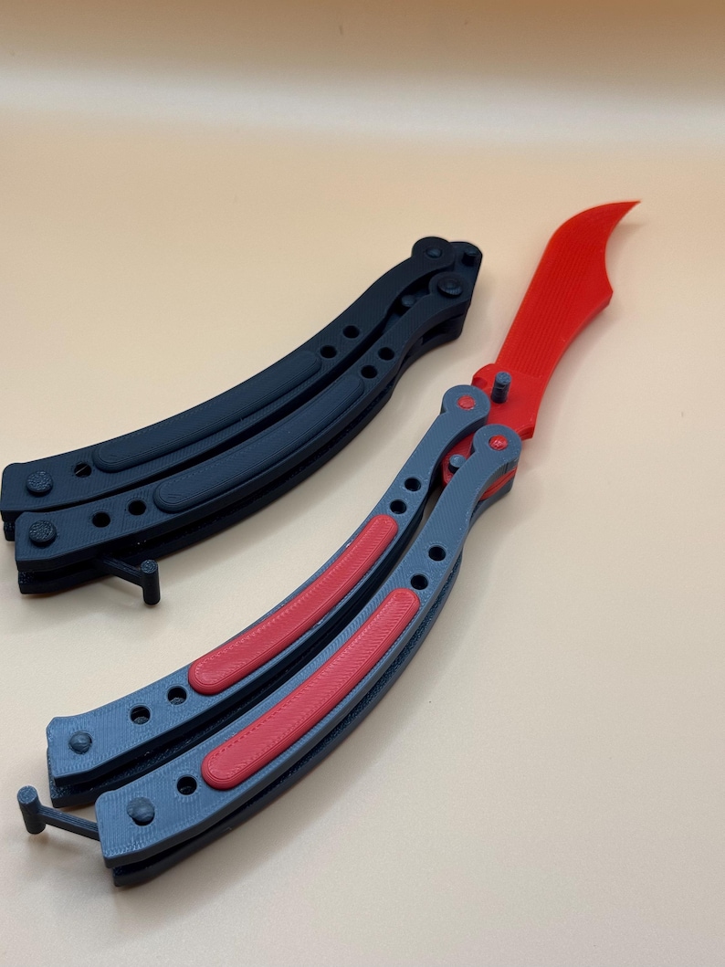 May include: Two butterfly knives, one black and one gray with red accents. The knives are folded and lying on a light brown surface.