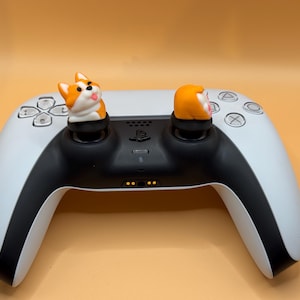 May include: A white and black PlayStation 5 DualSense controller with two orange and white dog-shaped joystick caps.