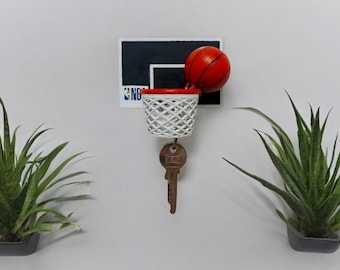 May include: A white basketball hoop with a red rim and a small orange basketball hanging from it. A silver key is attached to the hoop. The hoop is mounted on a white wall.
