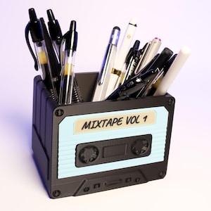 Peut inclure: Un organisateur de bureau en forme de cassette noire et bleue avec un design de cassette bleu clair et le texte "MIXTAPE VOL 1". L'organisateur est rempli de divers stylos et crayons.