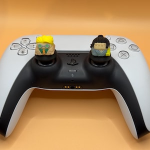 May include: A white and black PlayStation 5 controller with two custom character figures attached to the analog sticks. The figures are a yellow-haired character wearing glasses and a brown and green outfit, and a black-haired character wearing a blue and yellow outfit.