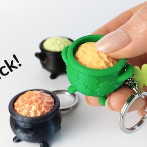 Può includere: Tre piccoli portachiavi a forma di calderone nero con un pentolino d'oro all'interno, di colore verde, arancione e giallo. Il portachiavi verde ha un ciondolo a forma di trifoglio. Il testo "Click!" è visibile nell'immagine.