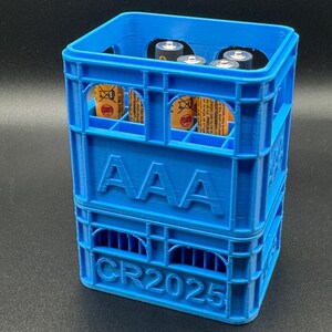 May include: A bright blue plastic crate, featuring the text "AAA" and "CR2025", containing several AAA batteries. The crate has a lattice design. The batteries are visible through the crate's openings. The crate is made of solid blue plastic.