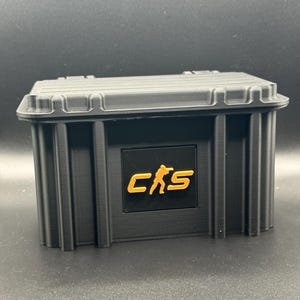 May include: Black rectangular storage box with a ridged lid and vertical side panels. The box features a black square with the gold letters "CKS" and a silhouette of a person.