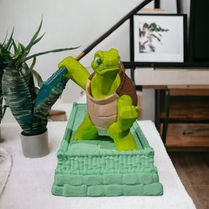 Puede incluir: Una figurita de tortuga verde sosteniendo una espada azul, de pie sobre una base de piedra verde. La tortuga tiene un caparazón marrón y un cuerpo verde.