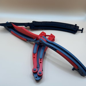 May include: Two butterfly knives, one red and black, the other black. The knives are folded and lying on a white surface.