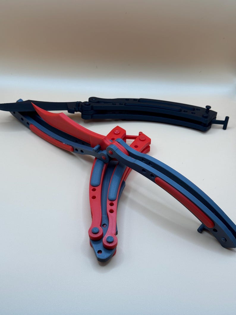 May include: Two butterfly knives, one red and black, the other black. The knives are folded and lying on a white surface.