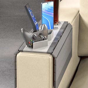 May include: A gray and white sofa armrest organizer with multiple compartments. It holds a remote control, a smartphone, a video game controller, and a glass of a dark beverage. The organizer is designed to fit over the arm of a sofa.