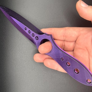 May include: A purple and black plastic knife with a finger hole in the handle. The knife has a textured surface and multiple holes in the handle.