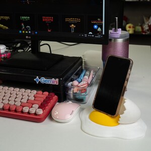 May include: A white and yellow phone stand shaped like a cracked egg with a yolk. A black smartphone is in the stand. A pink and red keyboard and a pink mouse are on a white desk.