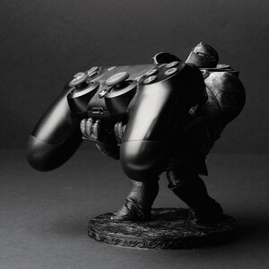 Puede incluir: Una figura impresa en 3D en negro y gris de una persona sosteniendo un mando de PlayStation negro. La figura está de pie sobre una base redonda con un diseño decorativo.