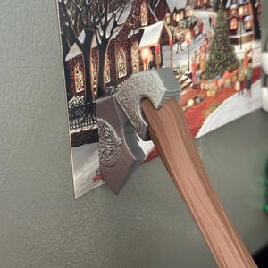 Könnte beinhalten: Ein dekorativer Magnet in Form einer Axt mit einem silberfarbenen Kopf und einem braunen Holzgriff. Die Axt hält eine Weihnachtskarte mit einer verschneiten Dorflandschaft mit Kirche und Weihnachtsbaum.