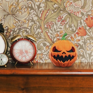 May include: A carved orange pumpkin with a scary face sits on a wooden shelf in front of a patterned wallpaper. Two vintage alarm clocks are on the shelf behind the pumpkin.