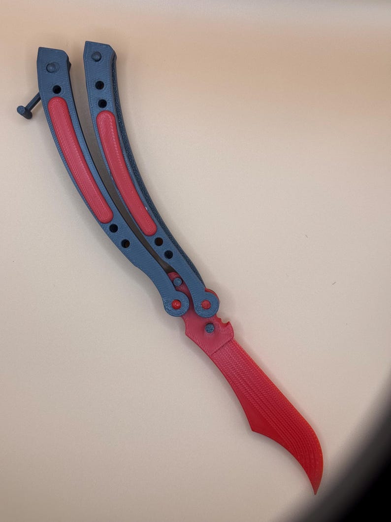 May include: A black and red butterfly knife with a curved blade. The handle is black with red accents.