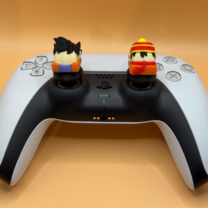 Könnte beinhalten: Ein weißer und schwarzer PlayStation 5-Controller mit zwei Figuren, die an den Analogsticks befestigt sind. Die Figur links ist ein schwarzhaariger Charakter mit einem orangefarbenen und violetten Outfit. Die Figur rechts ist ein Charakter mit einem gelben und roten Hut und einem roten Schal.