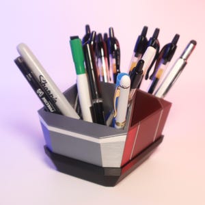May include: A multi-compartment desk organizer in gray, red, and black holds various pens and markers. The organizer has a geometric design with a white accent stripe. A green marker and a Sharpie pen are visible.