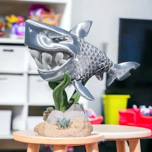Op de afbeelding: Een zilveren 3D-geprint haaienbeeldje met een tandenrij, gepositioneerd op een basis met groen zeewier en rotsen.
