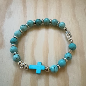 May include: A turquoise blue beaded bracelet with a silver cross charm and a silver charm of the Virgin Mary.