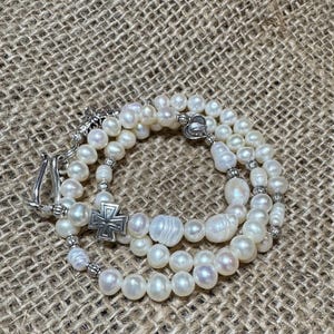 Puede incluir: Un collar de perlas blancas con un dije de cruz de plata y un cierre de plata. El collar está en capas y tiene un aspecto delicado.