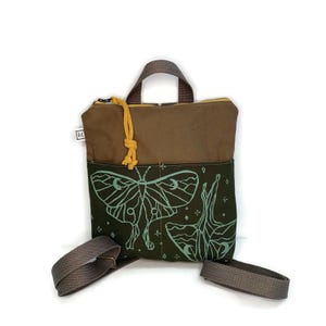 May include: A small brown canvas backpack with adjustable straps. The front pocket features a green and white print of two moths with moon and star details.