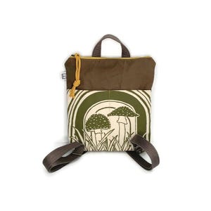 May include: A small backpack with a brown top and a green and white mushroom design on the front. The backpack has adjustable straps and a top handle. The zipper is yellow and the bag has a small tag on the side.