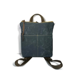 May include: A dark charcoal gray backpack made of waxed canvas featuring a top handle, two adjustable brown webbing shoulder straps, a zippered main compartment with a brown leather pull, and a front exterior pocket with yellow stitching.