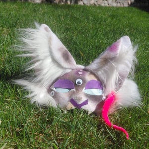 May include: A handmade, whimsical mask with a cat-like design. The mask features large, fluffy white ears with purple accents, purple eye coverings, and a pink, curly tail. The mask is lying on a bed of green grass.