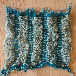 Può includere: Un tessuto intrecciato color verde acqua e beige con un motivo testurizzato e ondulato. Il tessuto ha un bordo sfrangiato.