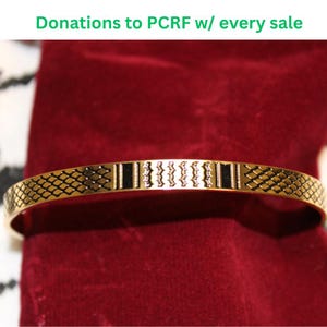 Może przedstawiać: Złota bransoletka z geometrycznym wzorem. Bransoletka ma powtarzający się wzór w kształcie rombów i pionowych pasków. Tekst "Donations to PCRF w/ every sale" jest widoczny u góry.