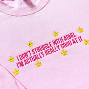 以下が含まれることがあります： ピンク色のクルーネックのスウェットシャツ。ピンク色の刺繍で「I DON'T STRUGGLE WITH ADHD, I'M ACTUALLY REALLY GOOD AT IT」と書かれています。黄色の星がテキストを囲んでいます。柔らかい素材で作られたスウェットシャツです。
