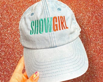 May include: Pale blue baseball cap featuring the embroidered text "SHOWGIRL" in green and orange. The cap has a curved peak and a classic six-panel construction. The hat is made from a soft, denim-style fabric.