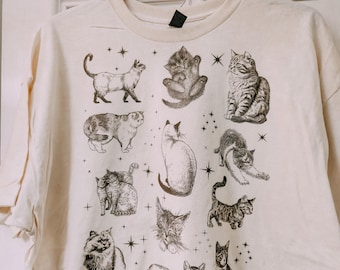 May include: Cream-colored t-shirt featuring a collection of cat illustrations in various poses. The drawings are in black and white, with star accents. The shirt is hanging on a black hanger with the brand name "Tu".