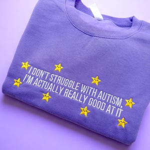 May include: A lavender sweatshirt with the embroidered text "I don't struggle with autism, I'm actually really good at it." Yellow star graphics are embroidered around the text. The sweatshirt is folded on a lavender surface.