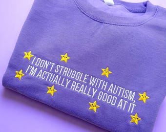 May include: A lavender sweatshirt with the embroidered text "I don't struggle with autism, I'm actually really good at it." Yellow star graphics are embroidered around the text. The sweatshirt is folded on a lavender surface.