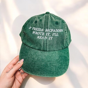 May include: Green baseball cap with white embroidered text: "IF FREIDA MCFADDEN WROTE IT, I'LL READ IT". The cap has a curved peak and is made of a soft, textured material. A hand is holding the cap.