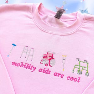 May include: A light pink crewneck sweatshirt featuring embroidered illustrations of a blue cane, a grey walker, a pink wheelchair, yellow crutches, and a green rollator. Below the icons, the text reads "mobility aids are cool" in pink.