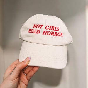May include: A cream-colored baseball cap with the embroidered text "HOT GIRLS READ HORROR" in red. The cap has a curved brim and a classic six-panel design, suitable for casual wear.