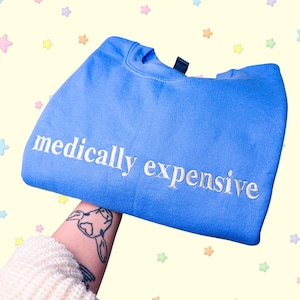 Op de afbeelding: Een felblauw sweatshirt met ronde hals en de tekst "medically expensive" geborduurd in witte, kleine schreefletters op de borst. Het kledingstuk heeft lange mouwen, een geribbelde halslijn en een standaard pasvorm, duidelijk in beeld gebracht.