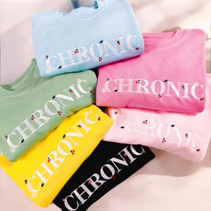 Op de afbeelding: Een stapel van zes sweatshirts in verschillende kleuren, elk met het woord "CHRONIC" geborduurd in wit met kleine rode kersen en witte bloemen. De sweatshirts zijn gevouwen en in een piramidevorm gerangschikt.