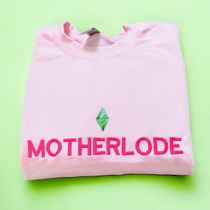 May include: A light pink sweatshirt with the word "MOTHERLODE" embroidered in hot pink, along with a green diamond graphic. The sweatshirt is folded neatly on a light green surface.