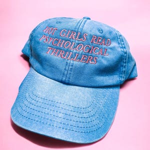 May include: A light blue denim baseball cap with the embroidered text "HOT GIRLS READ PSYCHOLOGICAL THRILLERS" in pink. The cap has a curved brim and a six-panel design, set against a pink background.
