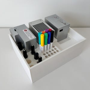 May include: A white wooden organizer box containing silver electronic devices, black cylindrical objects, and colorful markers. The markers include shades of blue, green, yellow, orange, purple, and black. The box is on a white surface.